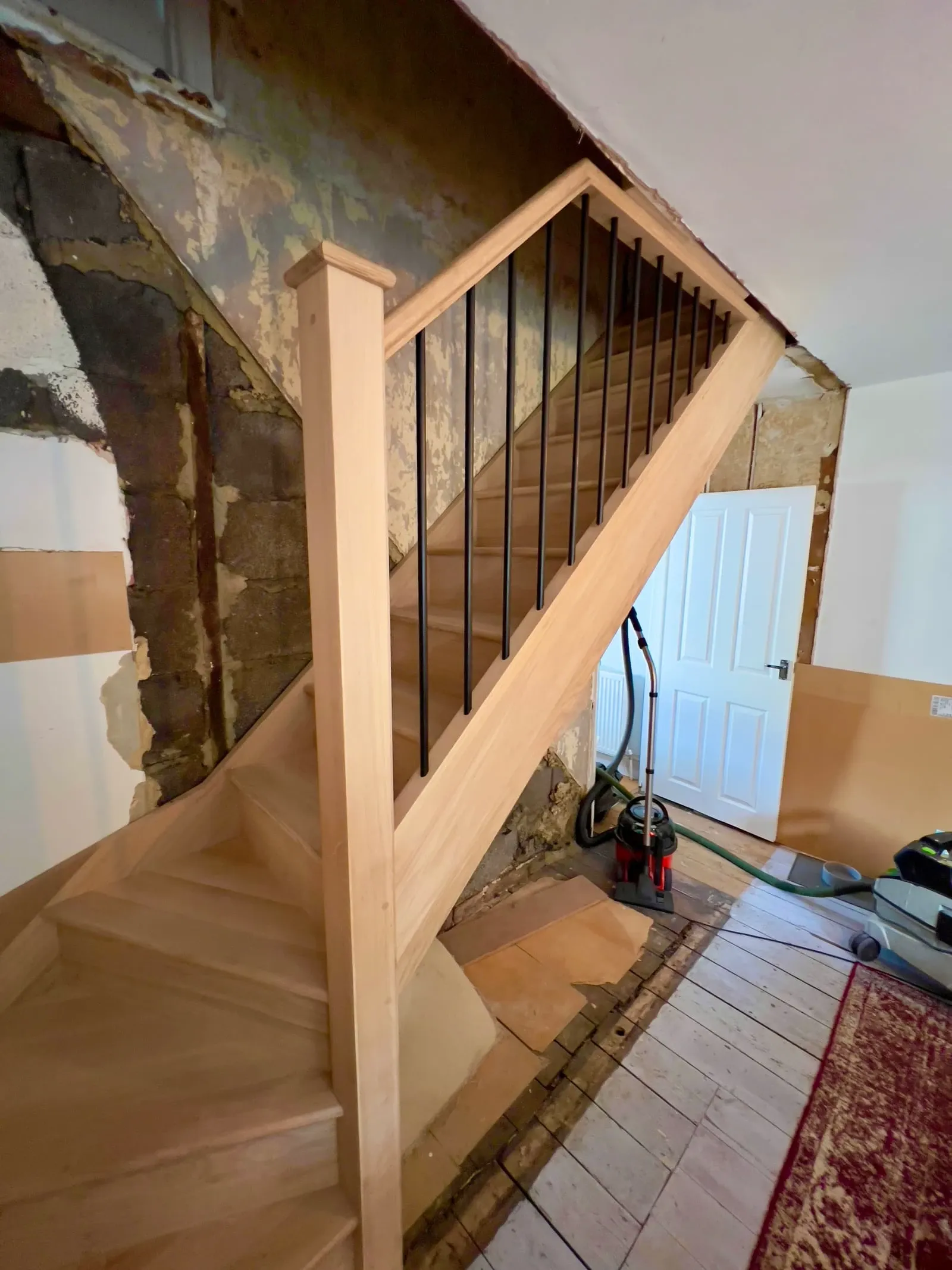 Side view of a completed oak staircase with sleek black spindles, set against an unfinished wall.