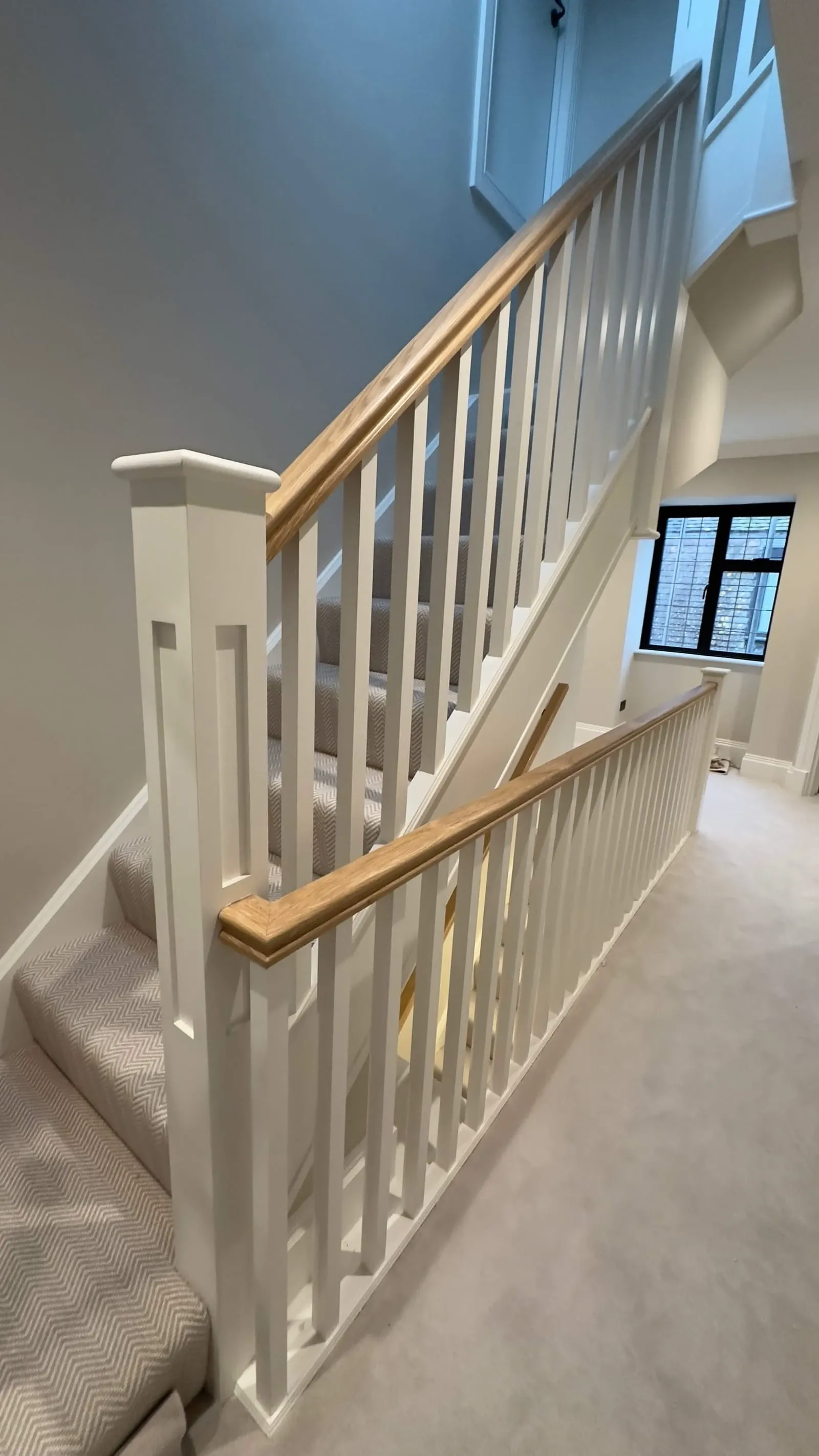 Modern staircase with white balustrades, oak handrails, and herringbone carpet, illuminated by natural light from a nearby window.