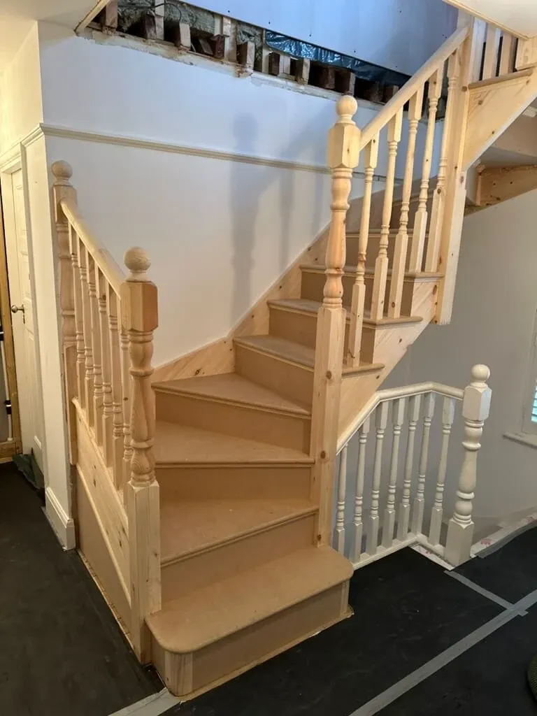 A newly installed wooden staircase with unfinished steps and spindles. The area above is under renovation with exposed beams.