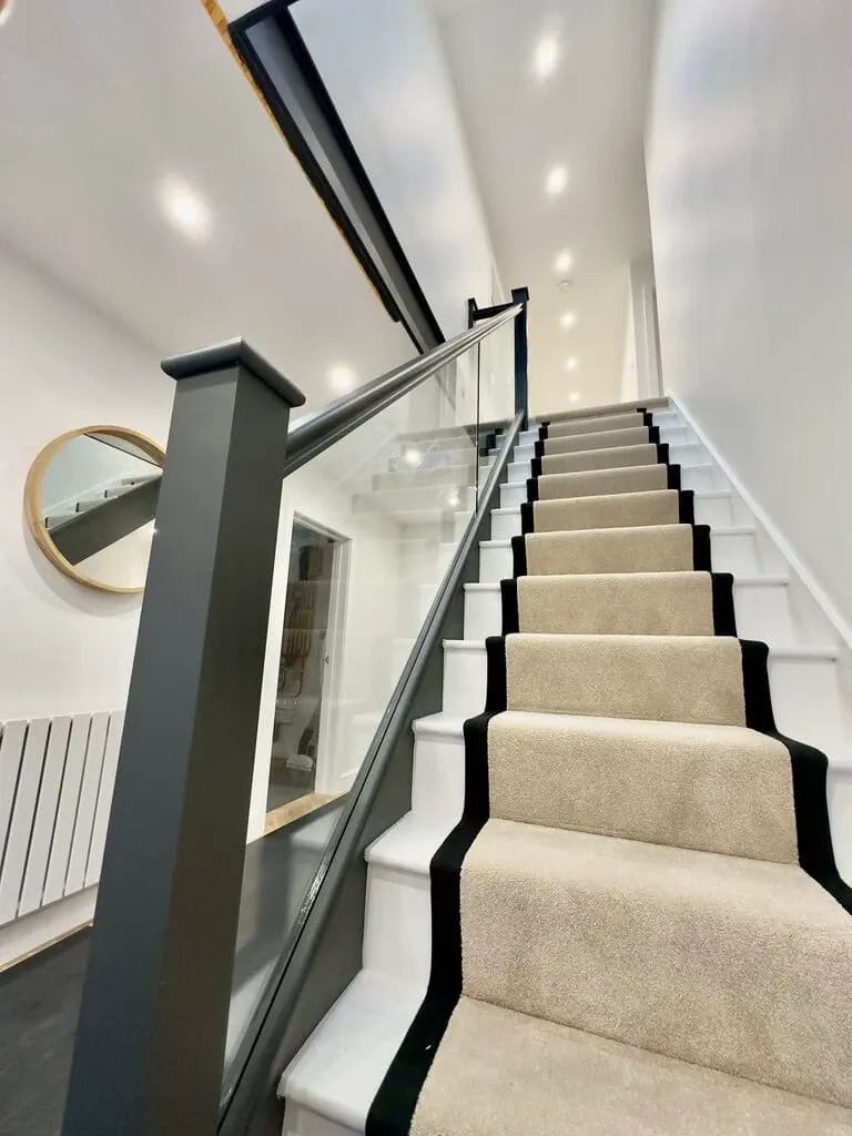 A modern staircase with beige carpeted steps bordered by black trim, glass balustrades, and dark grey frames. White walls and ceiling lights illuminate the space, with a round mirror and radiator on the left.