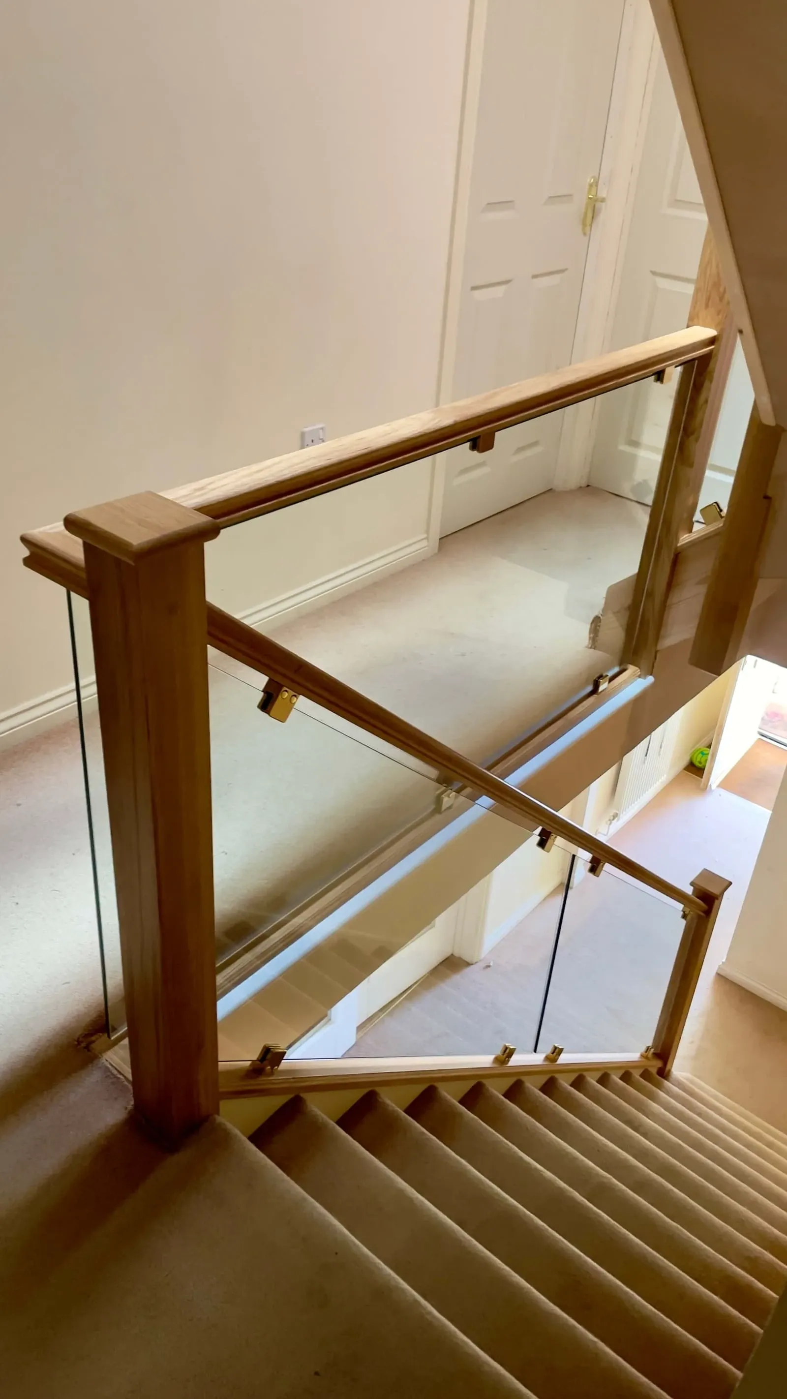 A modern staircase with beige carpeted steps bordered by black trim, glass balustrades, and dark grey frames. White walls and ceiling lights illuminate the space, with a round mirror and radiator on the left.