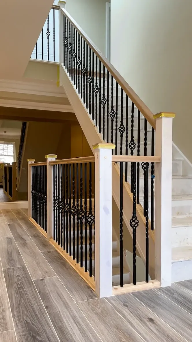 A staircase with unfinished white steps, black decorative balusters, and wooden handrails. Beige walls, a small window, and wood-effect tiled flooring are visible below.