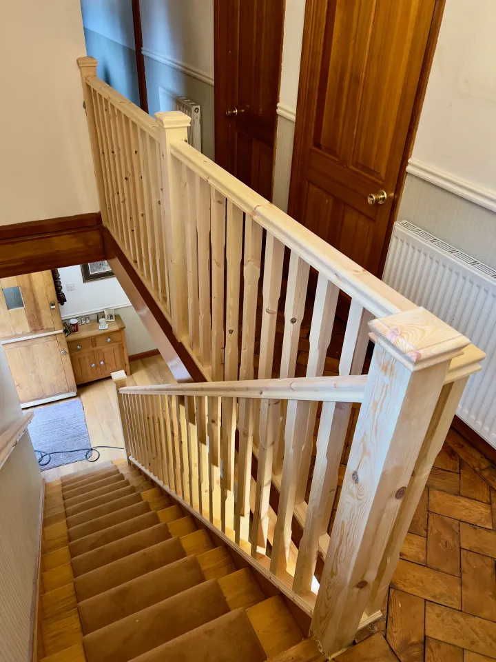 Modern wooden staircase with pine balustrades, handrails, and newel posts, located in a bright, carpeted hallway.