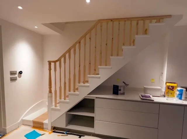 A modern staircase with wooden banisters, white steps, built-in storage, and a nearby worktop under bright ceiling lights.