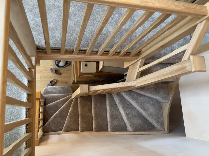 An oak staircase viewed from the top, with oak spindles and a grey carpet leading down to a wooden floor. 